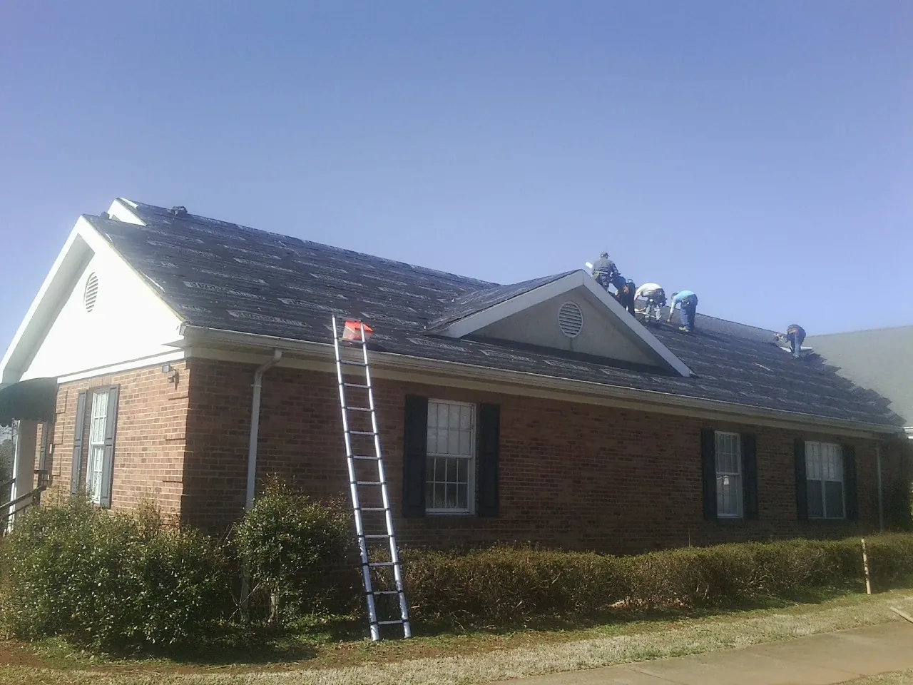 Crew tearing shingles off a brick ranch roof with a ladder set — Metal Roof Repair work by our crew in Castile