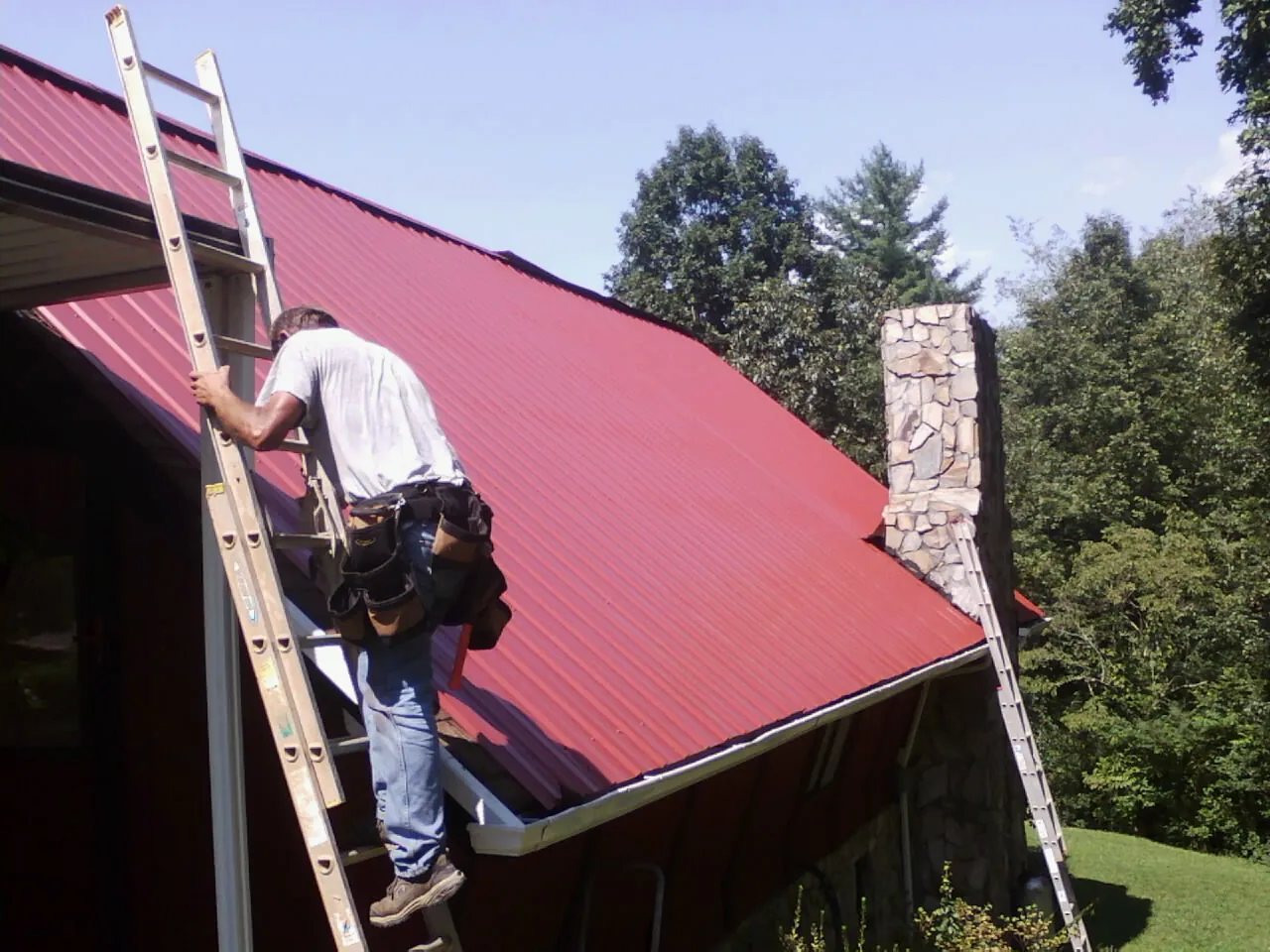 Roofer on a ladder beside a red metal roof and stone chimney — Soffit & Fascia Repair by Roofing Brothers in Castile