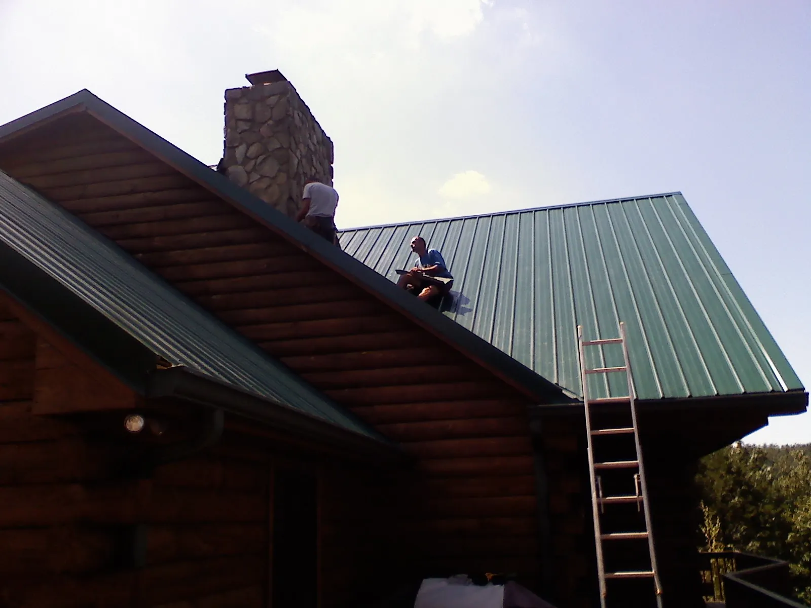 Roofers installing green metal panels beside a stone chimney on a log home in Castile
